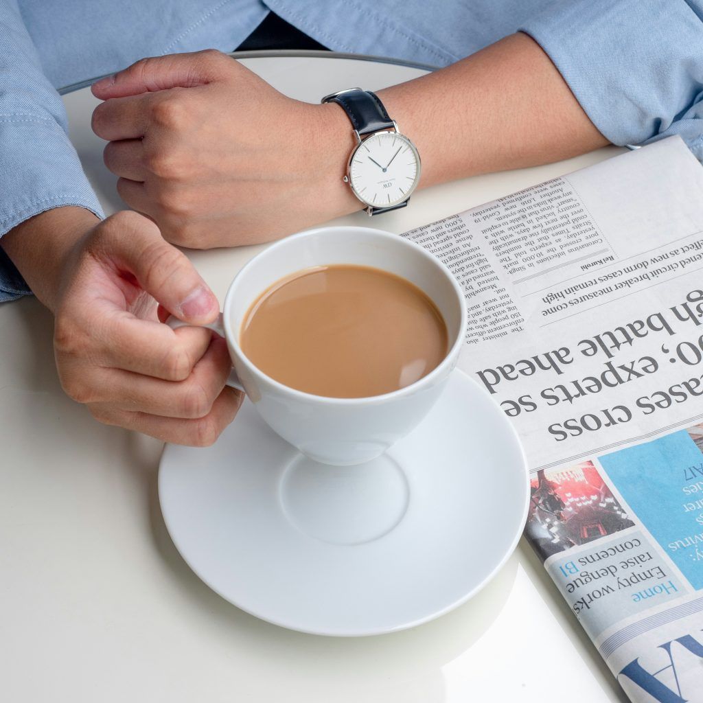 man wearing a minimalist watch holding a cup of coffee next to a folded newspaper